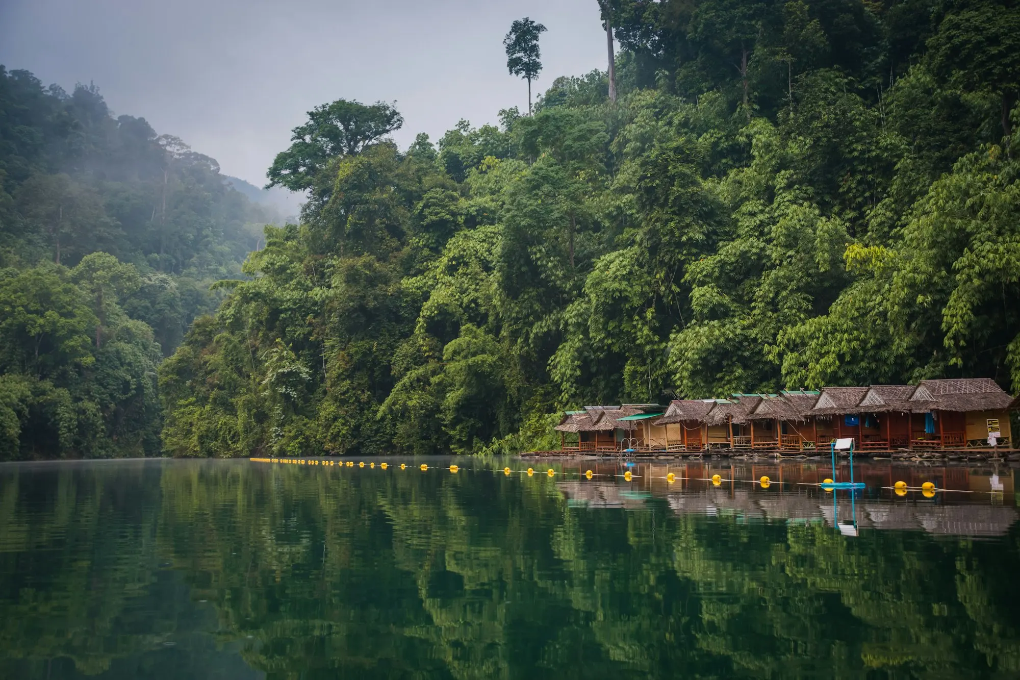 Wooden huts among green trees below limestone cliffs in Khlong Sok, Thailand